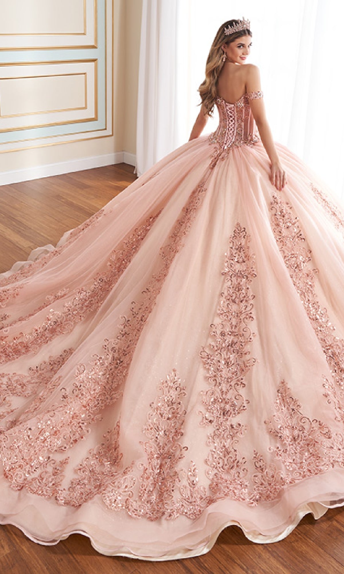 A young woman wearing the Quinceanera Dress PR30171 By Princesa, featuring intricate lace details and a tiara, stands on a wooden floor gazing over her shoulder.